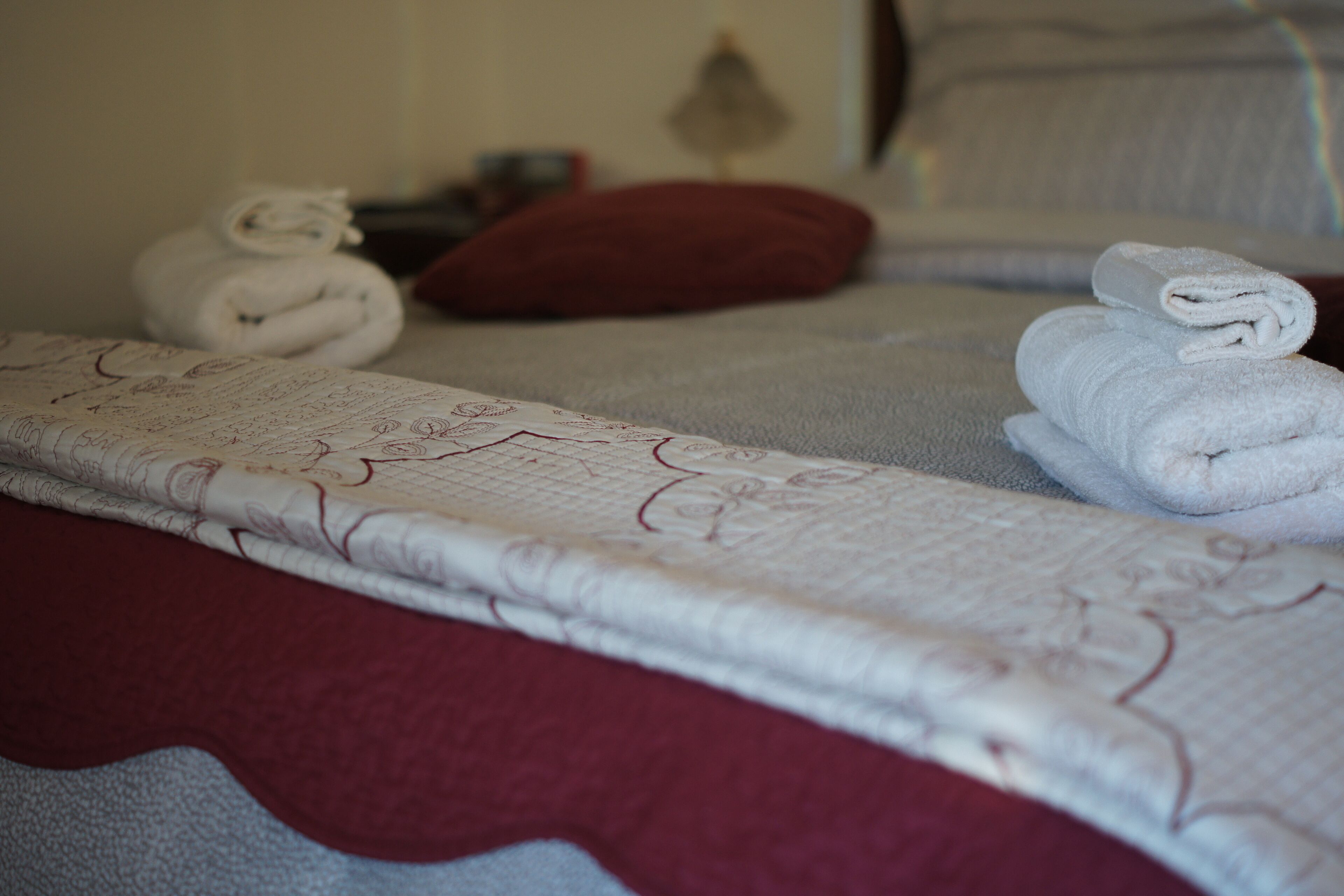 A neatly made bed with a patterned bedspread, two rolled towels, and a red pillow.