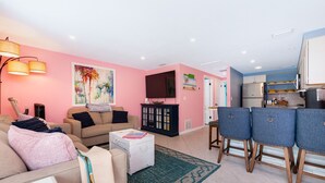 Una sala de estar con un sofá, una mesa de centro y un televisor. Hay una zona de cocina con refrigerador y alacenas.