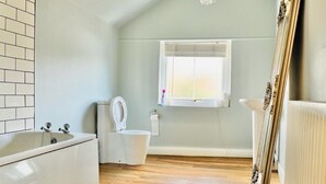 A bathroom with a bathtub, toilet, and a window with blinds. - Bodhi Tree Cottage - Grove Road Cottages (Stratford-upon-Avon)