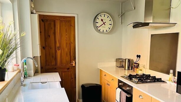 A kitchen with wooden cabinets, a white countertop, a stove, a sink, and a clock on the wall. - Bodhi Tree Cottage - Grove Road Cottages (Stratford-upon-Avon)