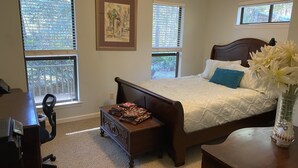 A bedroom with a bed, a desk, and a chair. There are windows with blinds, a framed picture on the wall, and a vase with flowers on the bedside table.