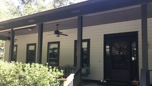 A covered porch with a black door, white siding, and a ceiling fan.