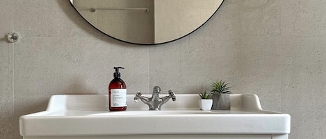 Ein Badezimmer mit weißem Waschtisch, einem runden Spiegel, einem Wasserhahn, einer Seifenspender und einem kleinen Blumentopf.