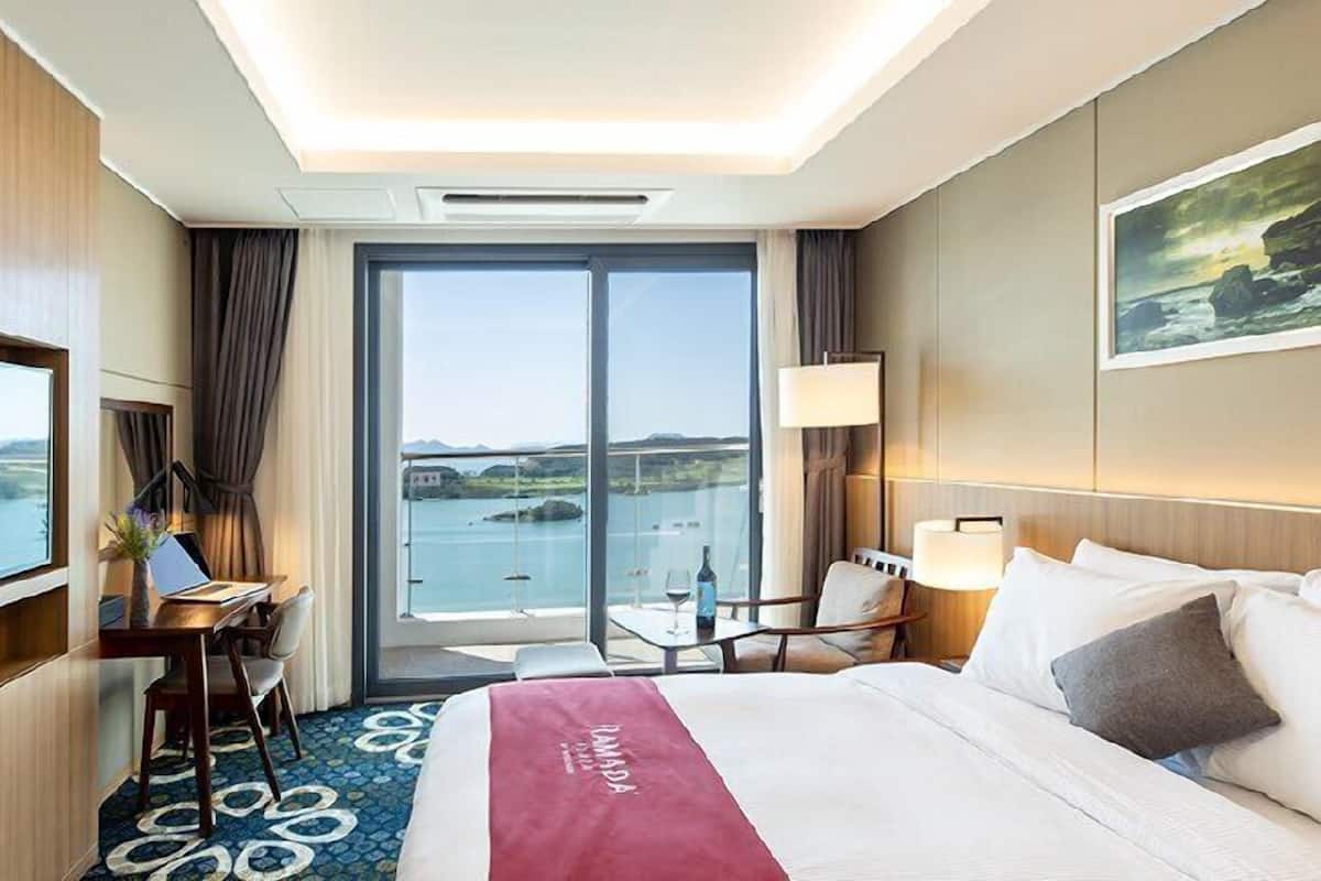 A hotel room with a large bed, a desk with a chair, a laptop, and a view of the sea through a large window.