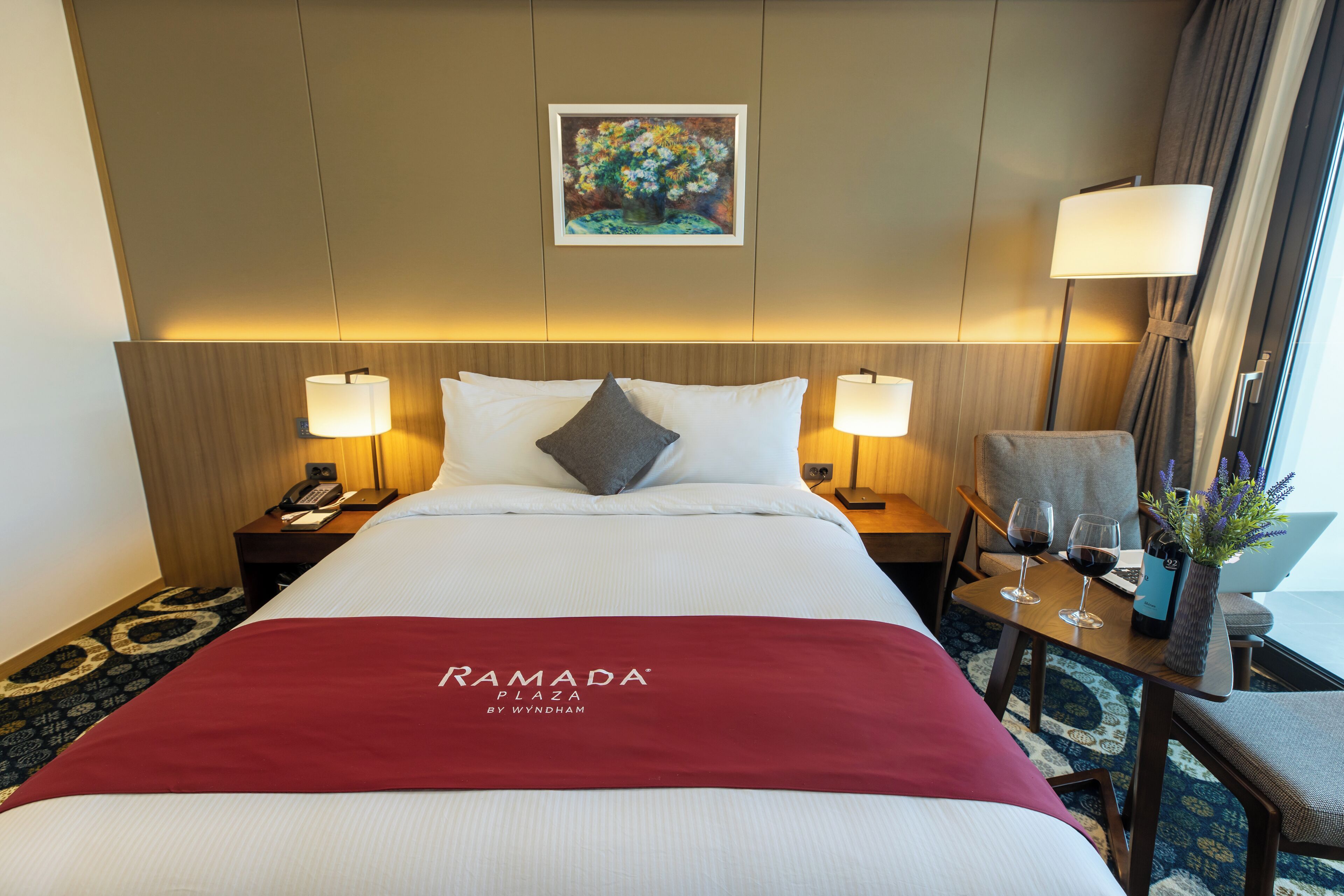 Una habitación de hotel con una cama, dos lámparas de noche, una mesita de noche, una silla, una pequeña mesa con dos copas de vino y un cuadro en la pared.