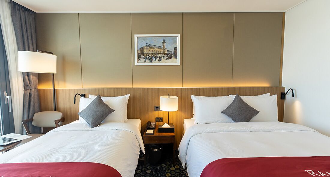 A hotel room with two beds, a desk, a chair, a lamp, a picture on the wall, and a trash bin.
