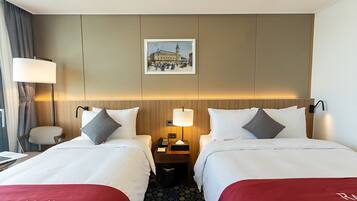 A hotel room with two beds, a desk, a chair, a lamp, a picture on the wall, and a trash bin.