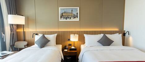 A hotel room with two beds, a desk, a chair, a lamp, a picture on the wall, and a trash bin.