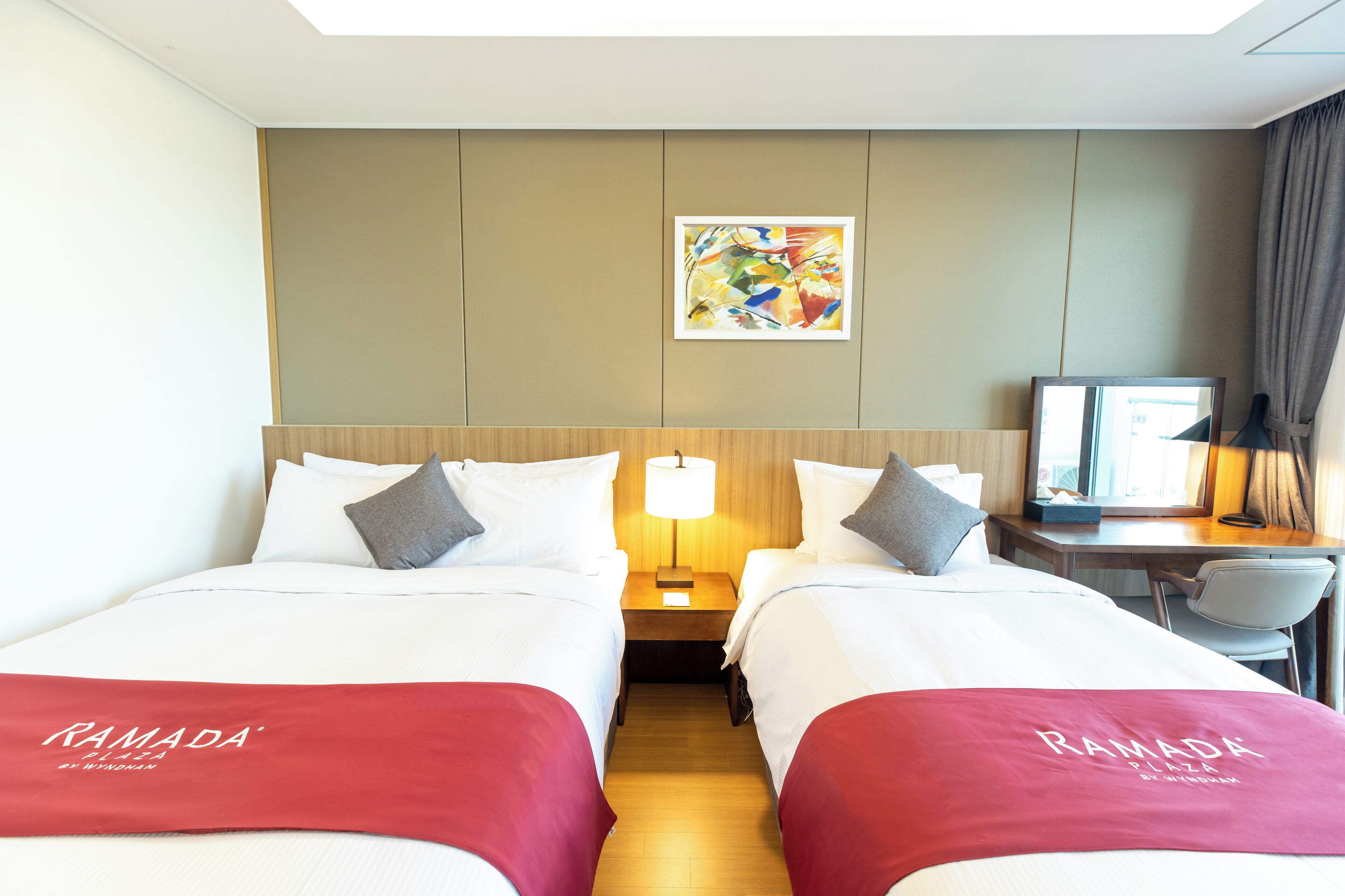 A hotel room with two beds, a desk, a chair, a lamp, and a painting on the wall.