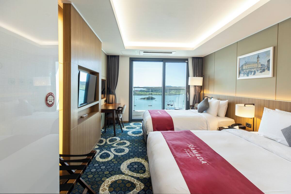 A hotel room with two beds, a desk, a TV, and a large window with a view of the water.