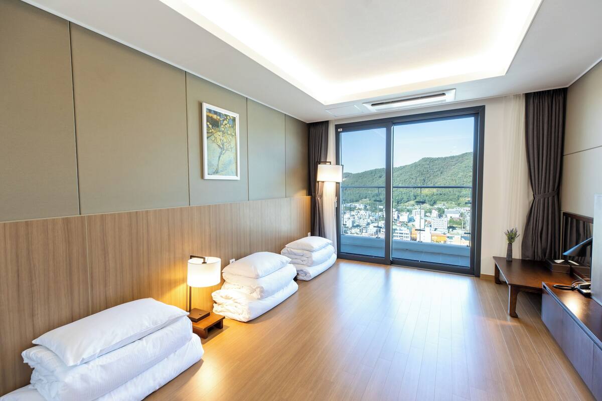 A hotel room with a large window, a bed with white linens, a bedside table with a lamp, and a view of a cityscape.