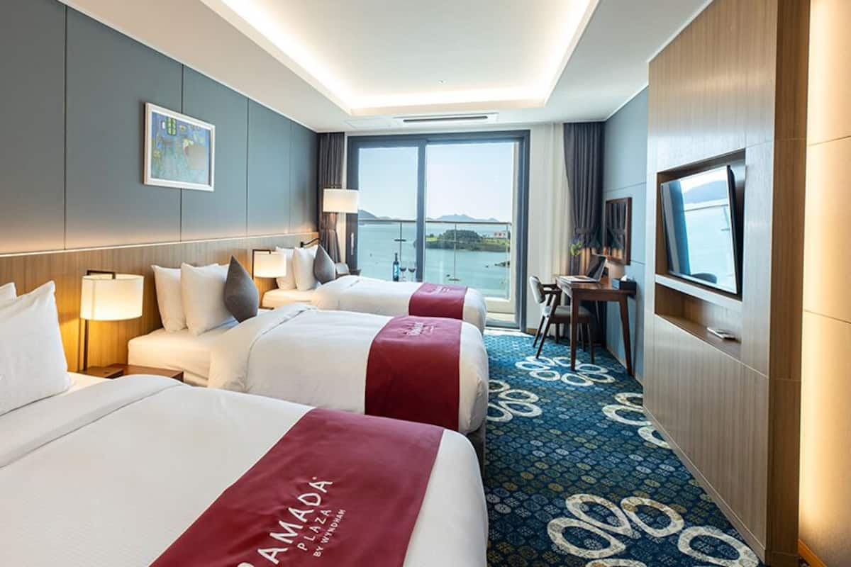 A hotel room with three beds, a desk, a TV, and a large window offering a view of the water.