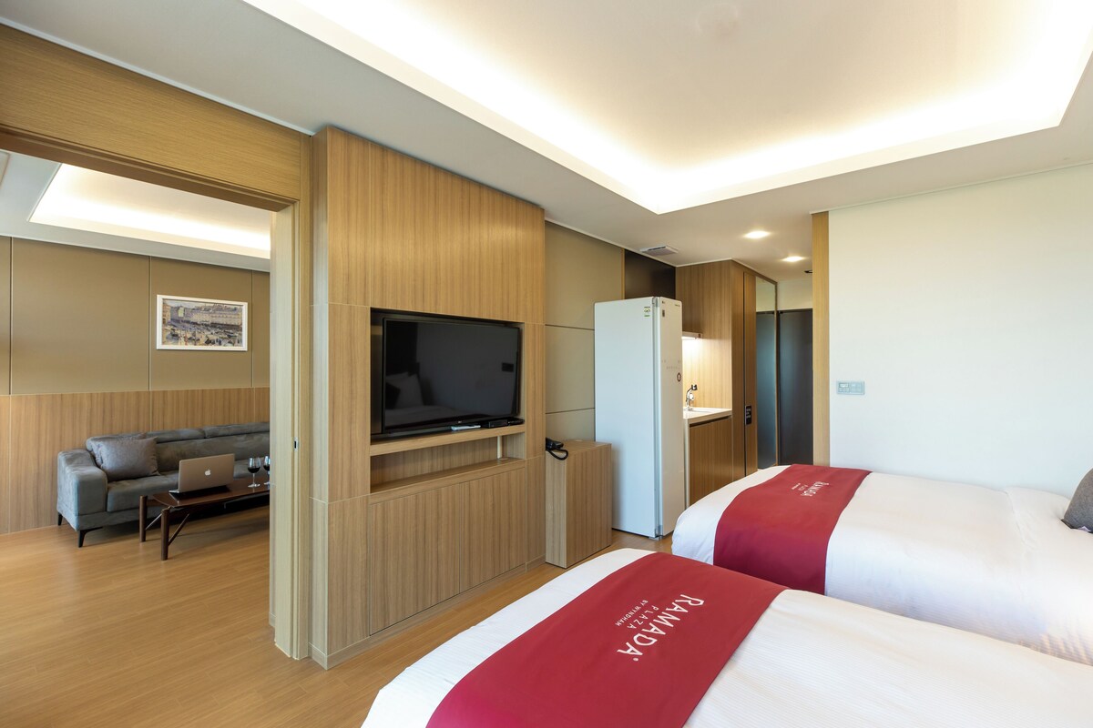 Una habitación de hotel con cama, televisor, escritorio con una computadora portátil, sofá y refrigerador.