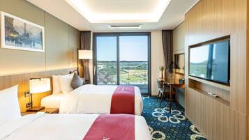 A hotel room with two beds, a TV, a desk, and a large window offering a view of the landscape.