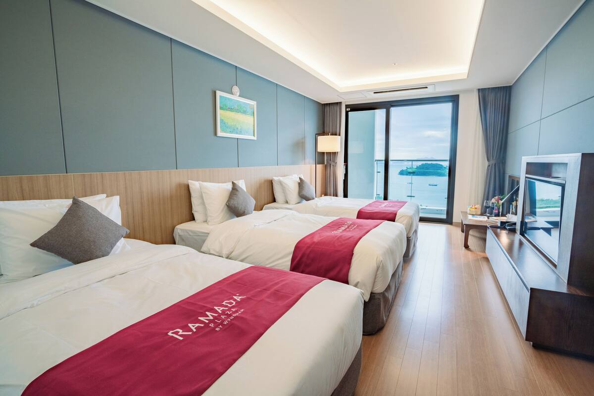 A hotel room with three beds, a TV, a desk, and a large window with a view of the sea.
