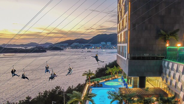 Vista de una playa con gente haciendo tirolesa, un área de piscina con palmeras y un edificio moderno con balcones.
