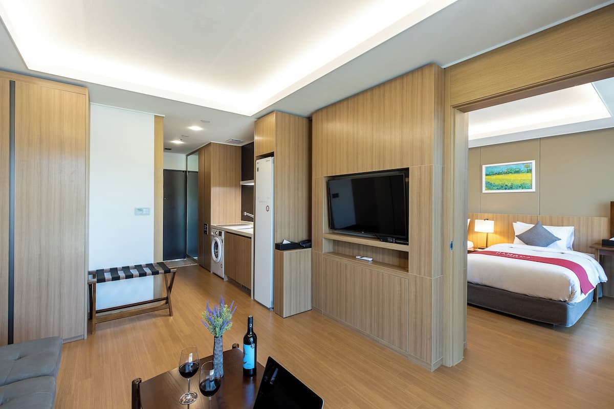 A modern hotel room with a bed, a TV, a dining area with a table and chairs, a sofa, and a small kitchen area with a washing machine.