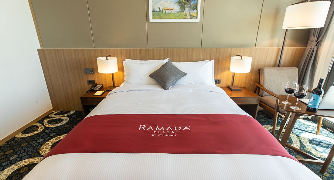 A hotel room with a bed, two bedside lamps, a chair, a small table with wine glasses and a bottle, and a red blanket with the hotel name.