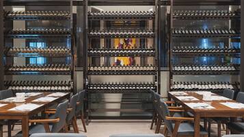 A wine cellar with rows of wine bottles on shelves, a long table with chairs, and a large painting on the back wall.