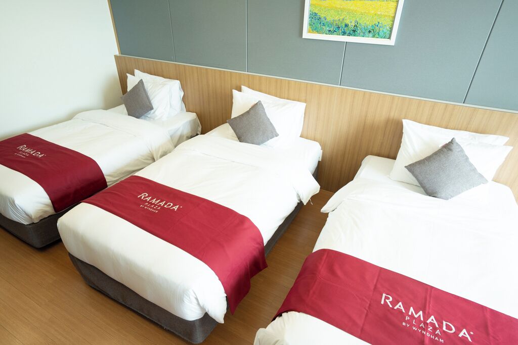 Tres camas individuales con ropa blanca y cabeceros rojos, cada una con una almohada y una manta roja con la marca 'RAMADA'.