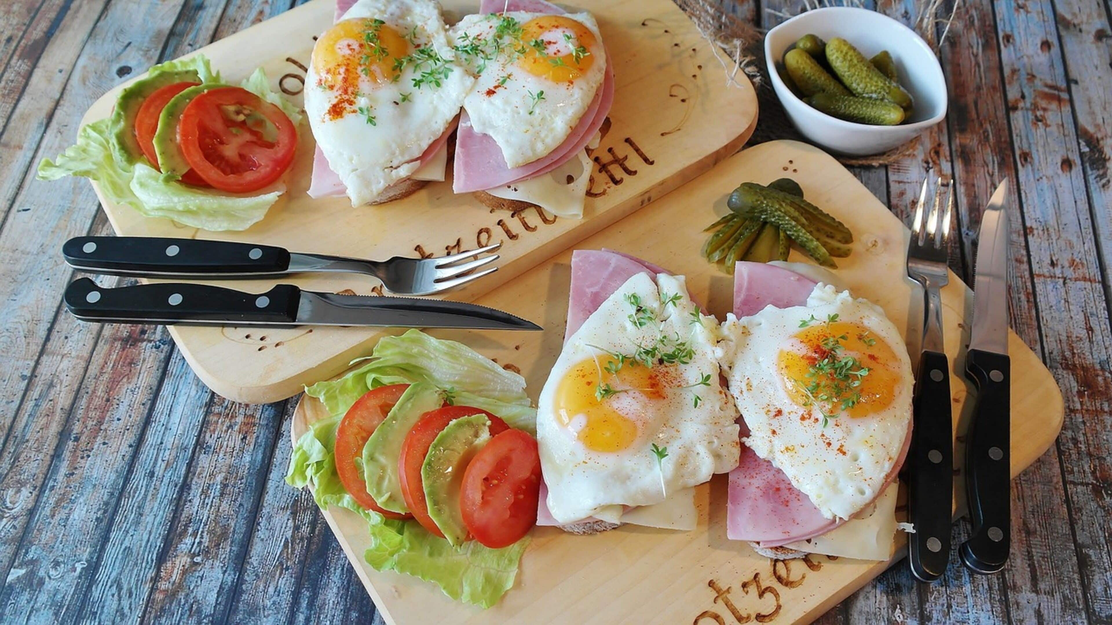 A wooden cutting board with two sandwiches, each with a fried egg, lettuce, tomato, and ham. There are also pickles and asparagus in a small bowl.