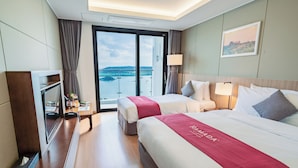 Habitación de hotel con dos camas, televisión, escritorio con lámpara y vista al mar.