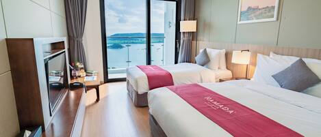 Habitación de hotel con dos camas, televisión, escritorio con lámpara y vista al mar.