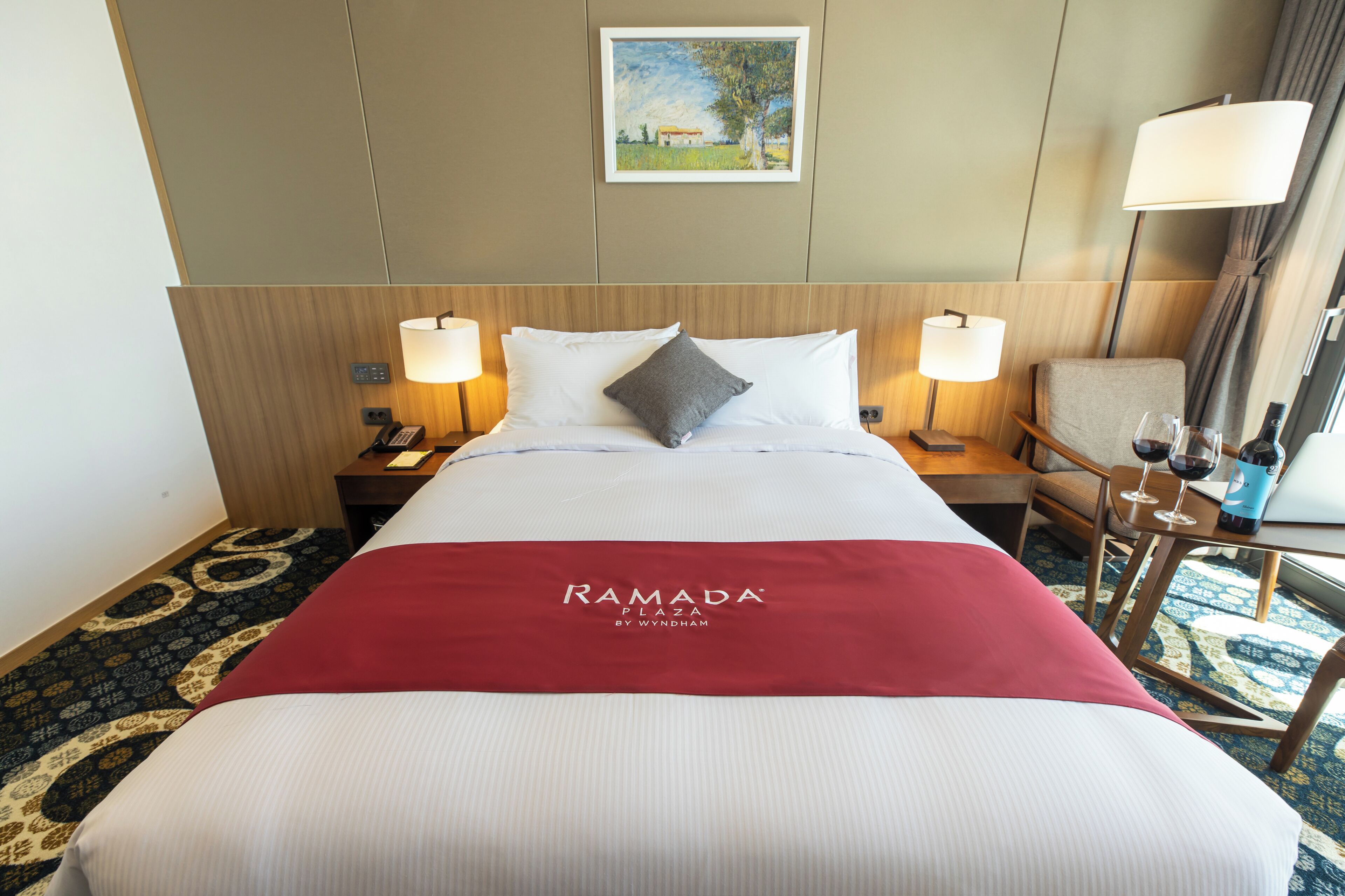 A hotel room with a large bed, two bedside tables with lamps, a chair, and a small table with wine glasses.
