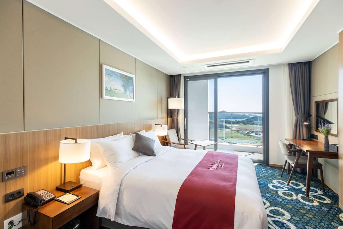 A hotel room with a large bed, bedside table with a lamp, a desk with a chair, a large window with a view, and a painting on the wall.
