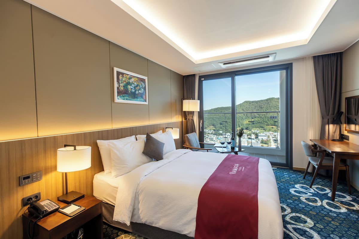 A hotel room with a large bed, a desk, a chair, a phone, and a window with a city view.
