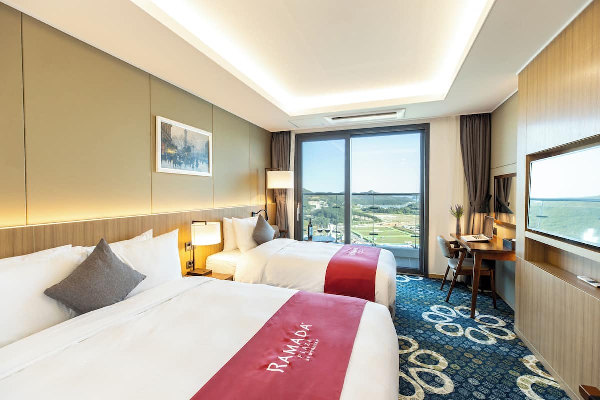 A hotel room with two beds, a desk, a chair, a TV, and a large window with a view.
