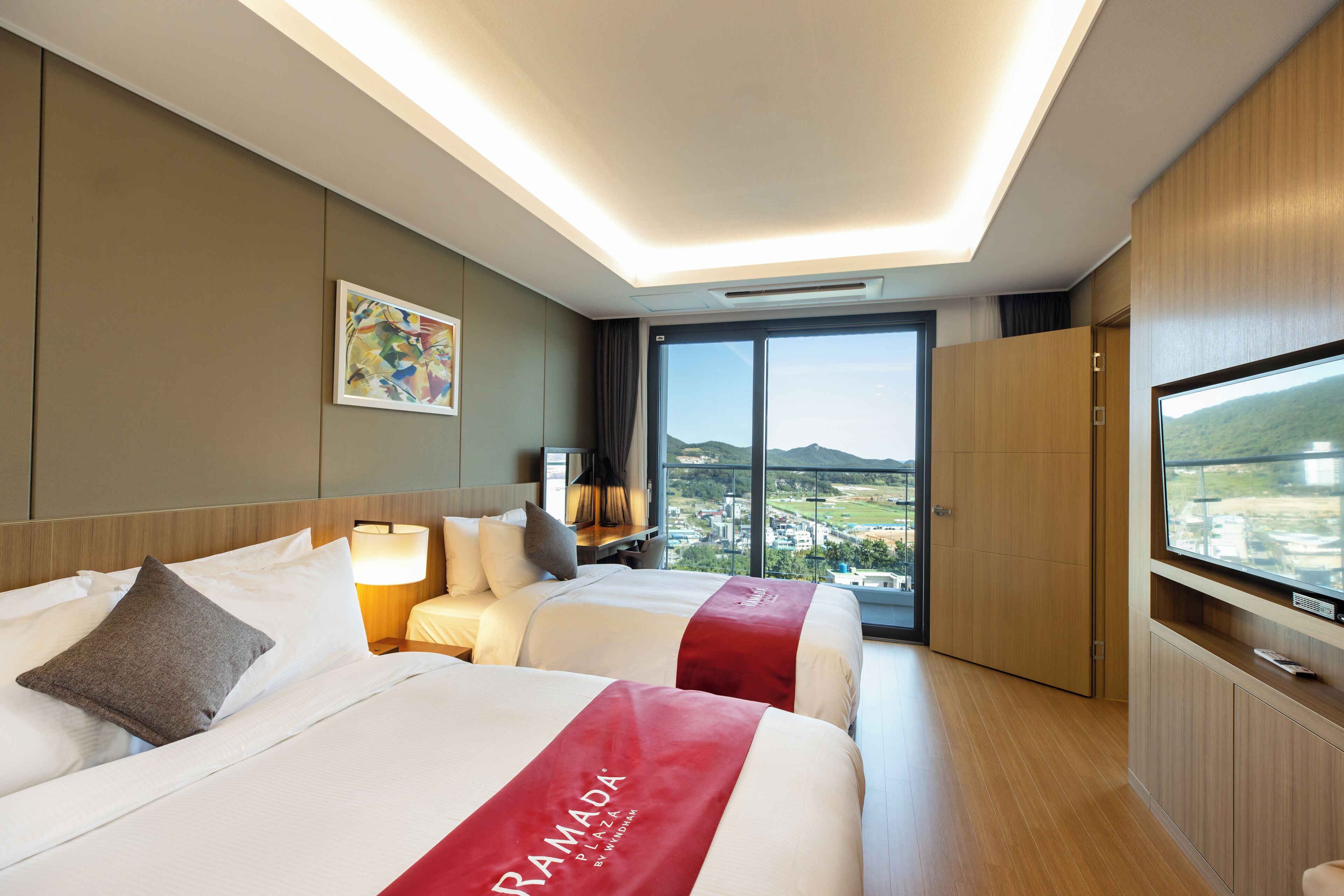 A hotel room with two beds, a TV, a painting, and a view of the city.