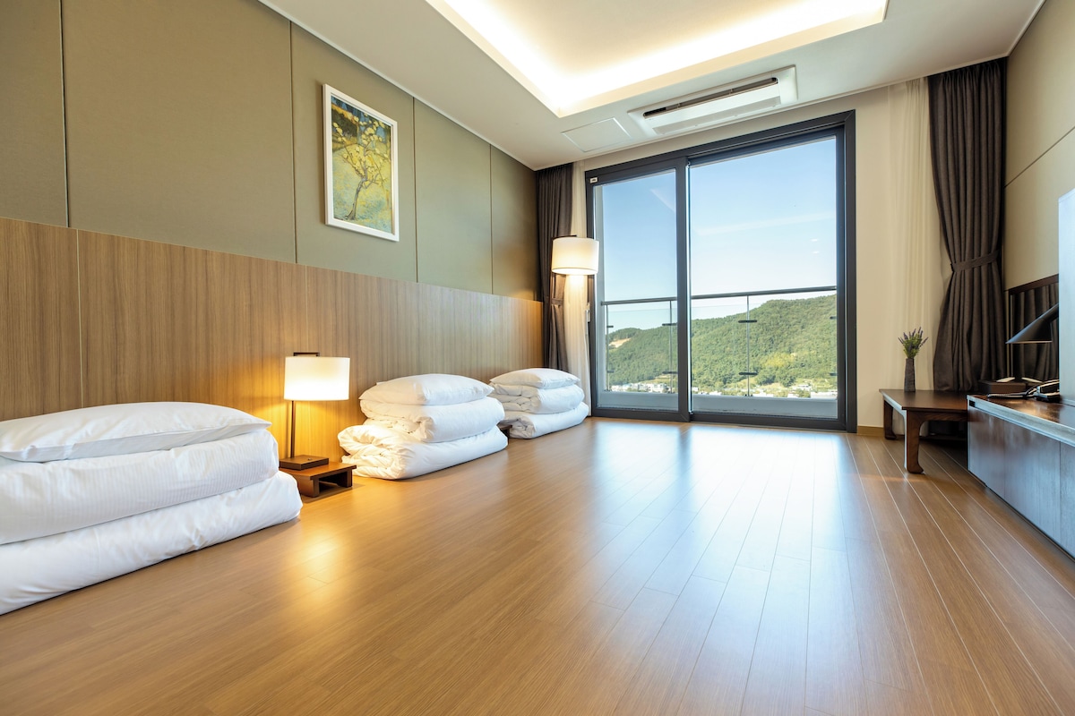 Habitación de hotel con una cama grande, un escritorio, una lámpara y vistas a una colina a través de un ventanal amplio.