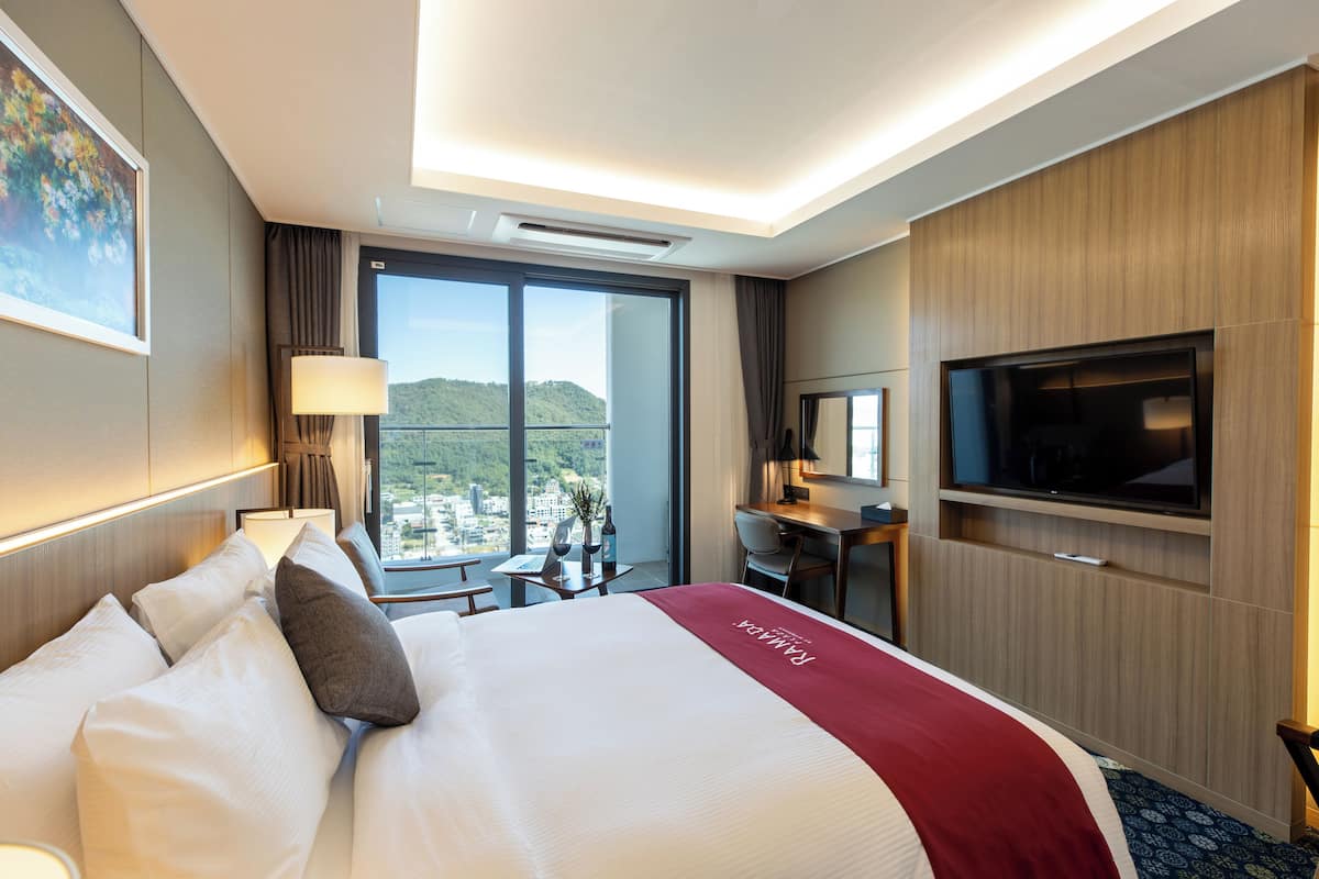 A hotel room with a large bed, a desk, a TV, and a view of the city.
