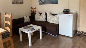 Una sala de estar con un sofá seccional marrón, una mesa de centro blanca, una silla de madera y un refrigerador blanco.