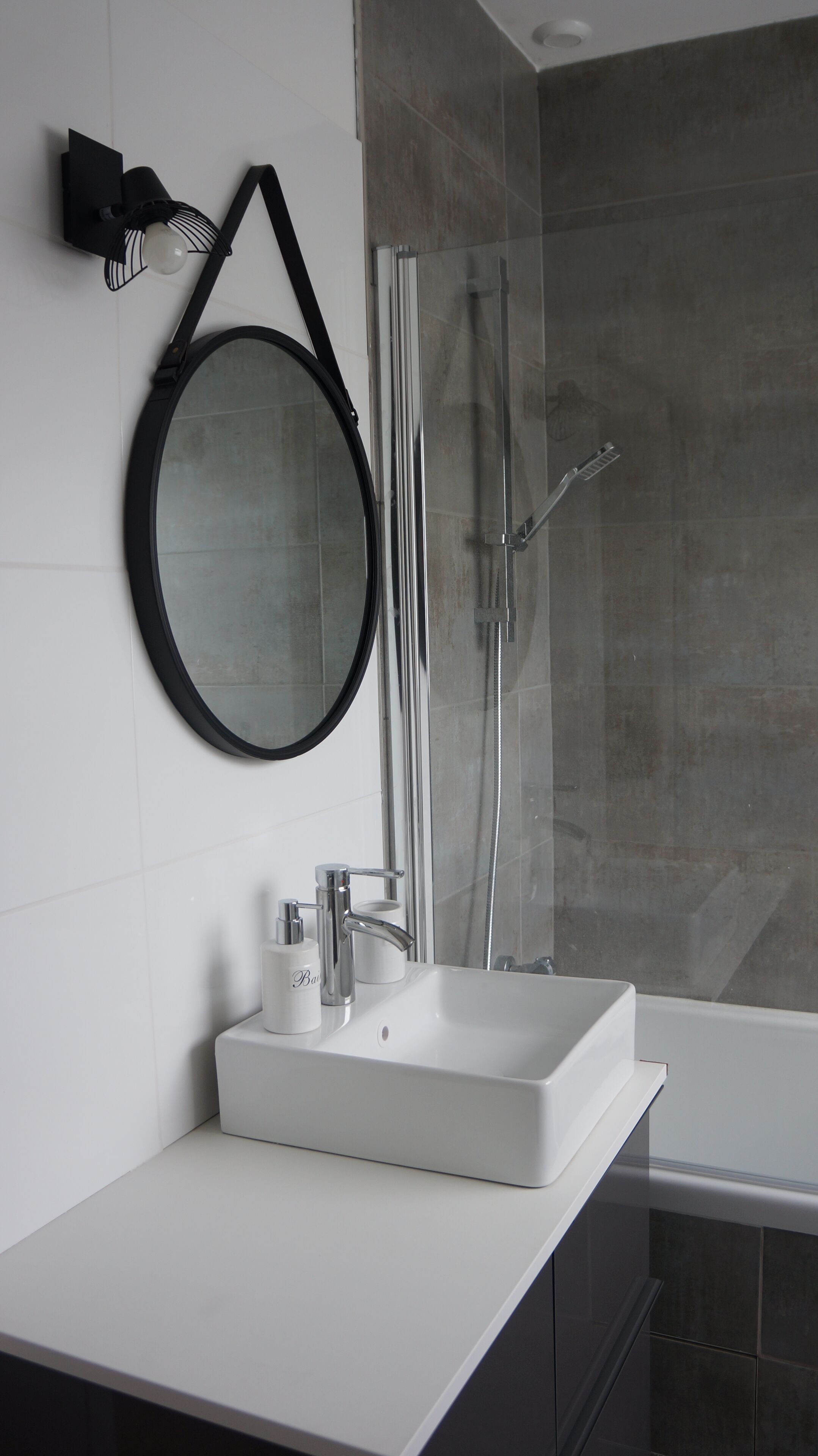 Un baño moderno con un lavamanos blanco, un espejo redondo y una ducha con mampara de vidrio.