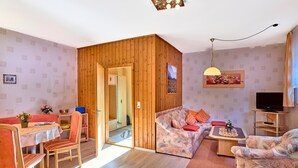 A cozy living room with a wooden partition, a dining area with a table and chairs, a sofa, a TV, and a small table with decorative items. - Ferienwohnungen Wirth (Bodenmais)