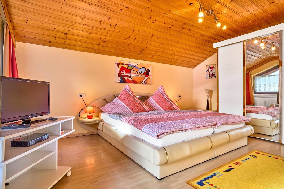 A bedroom with a wooden ceiling, a bed with patterned bedding, a TV on a stand, and artwork on the wall.