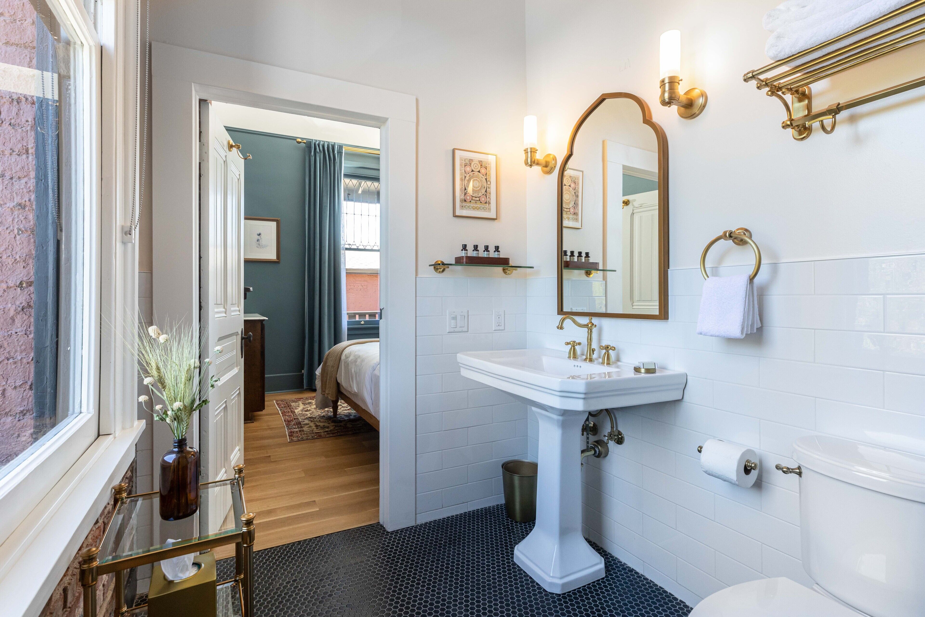 Ein Badezimmer mit einem weißen Waschbecken, einem Spiegel und einer Toilette. Ein Fenster bietet Blick auf einen Raum mit einem Bett und einem weiteren Fenster.