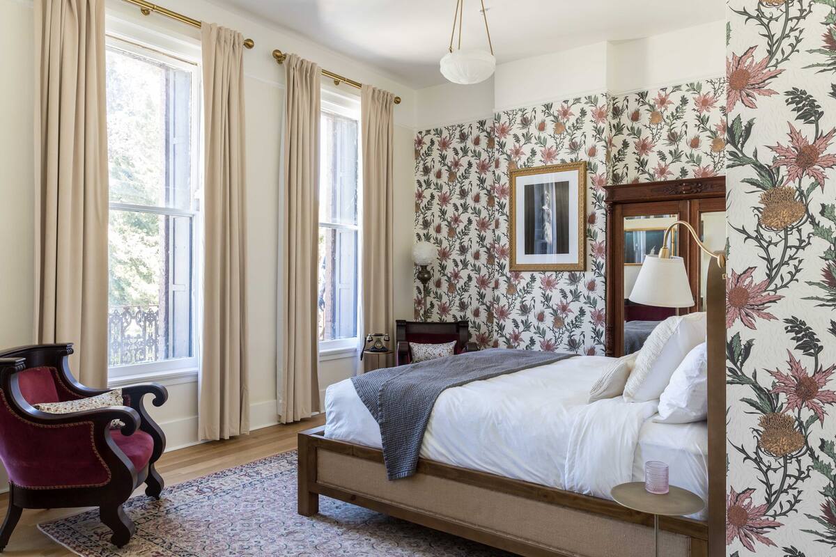 A bedroom with a bed, a chair, a nightstand, a lamp, and floral wallpaper.