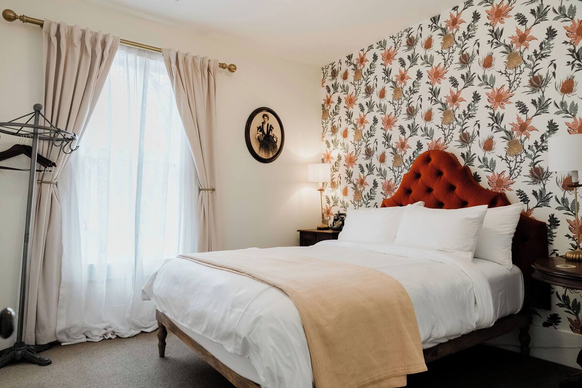 A neatly made bed with a red headboard, a wooden nightstand with a lamp, and a floral wallpaper.