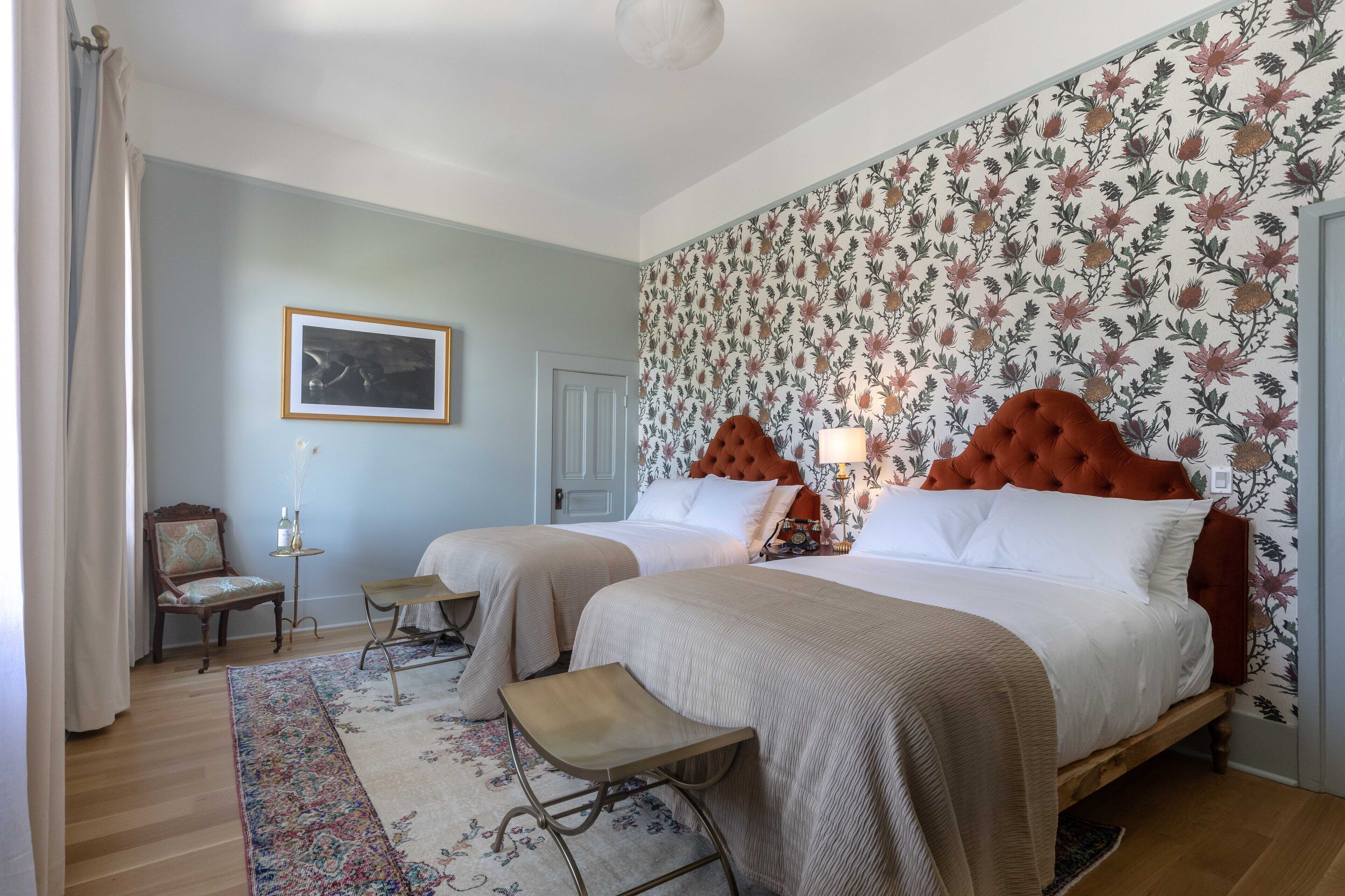 Une chambre d’hôtel avec deux lits, un papier peint à motifs floraux, un tableau encadré au mur, une petite table et une chaise.