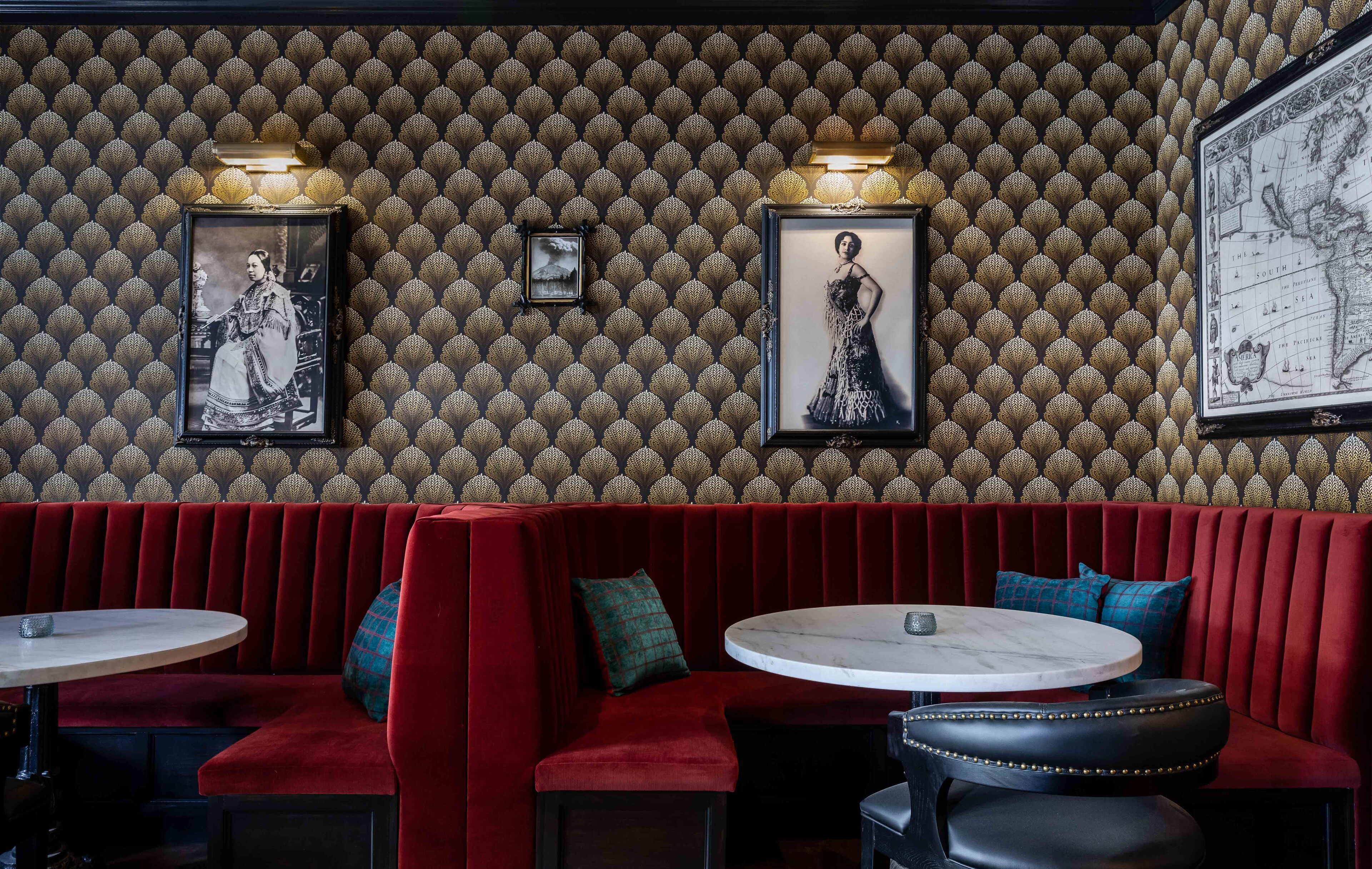A restaurant interior with red velvet booths, round tables, and patterned wallpaper.
