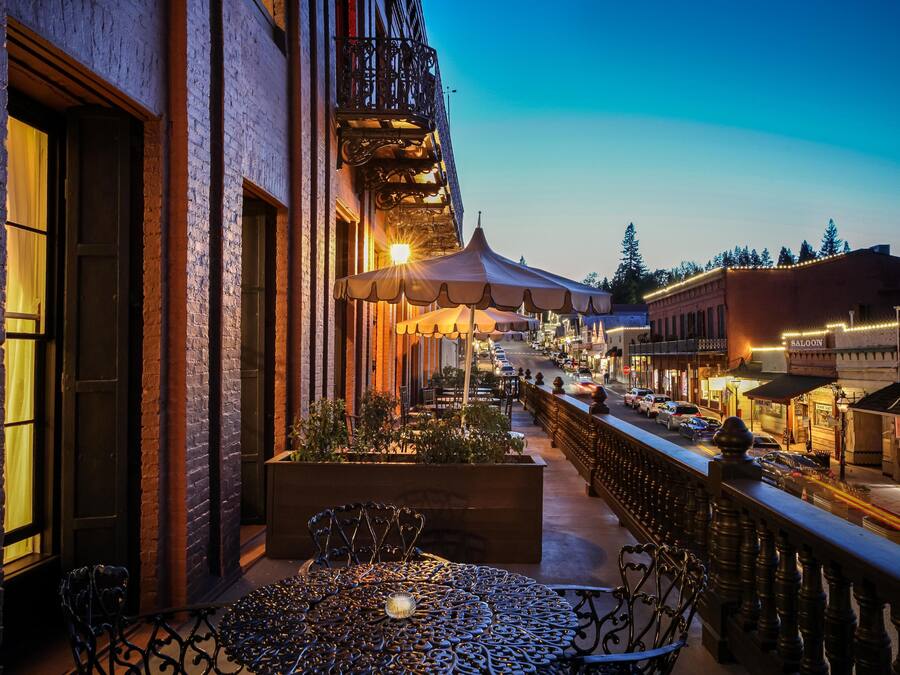 Eine Terrass mit schmiedeeisernem Tisch und Stühlen, ein Backsteinhaus mit Balkon und eine Straßenansicht mit Autos und Geschäften.