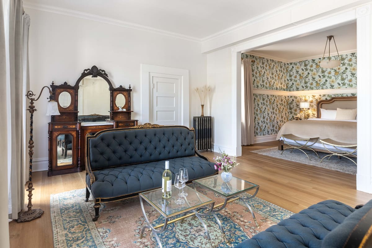 A room with a dark blue sofa, a glass coffee table, a wooden dresser with a mirror, and a bed with a floral wallpaper.