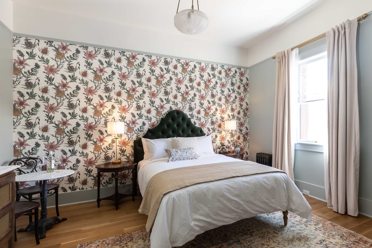 A bedroom with a floral wallpaper, a green tufted headboard, a white bedspread, and a round table with chairs.