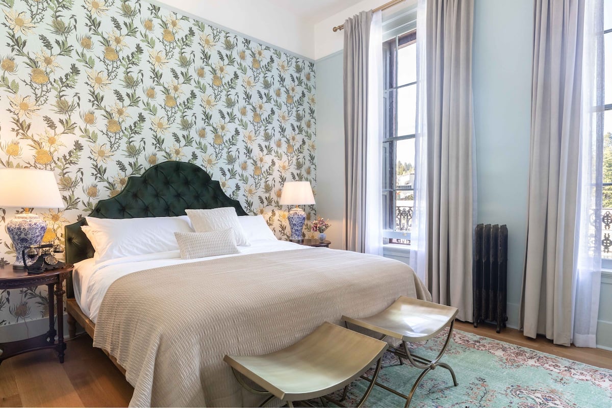 A neatly made bed with a tufted headboard, two bedside tables with lamps, a bench at the foot of the bed, and a floral-patterned wallpaper.