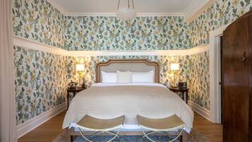 A bedroom with floral wallpaper, a large bed with a wooden headboard, two bedside tables with lamps, and a wooden wardrobe.