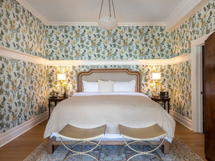 A bedroom with floral wallpaper, a large bed with a wooden headboard, two bedside tables with lamps, and a wooden wardrobe.