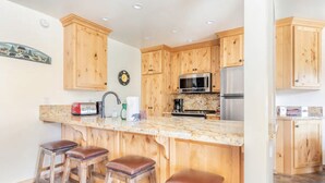 Una cocina con armarios de madera, encimera de mármol, microondas, refrigerador y tres taburetes.